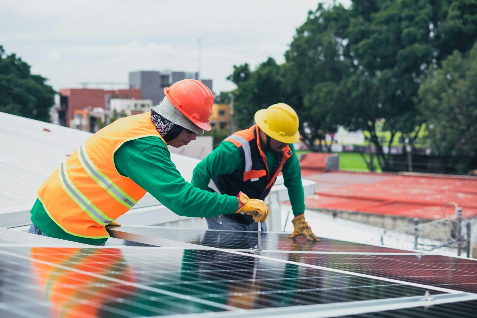 pexels-cristian-rojas-8853536 Two workers in safety gear install solar panels on a rooftop, with trees and buildings in the background.
