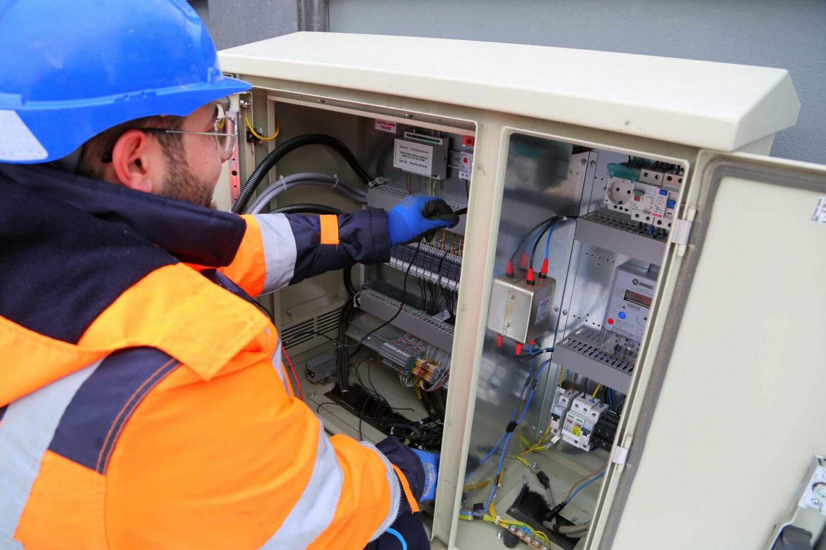 pexels-fatihyurtman-17842832 A worker in a blue hard hat and orange jacket inspects and adjusts controls inside an open electrical cabinet.